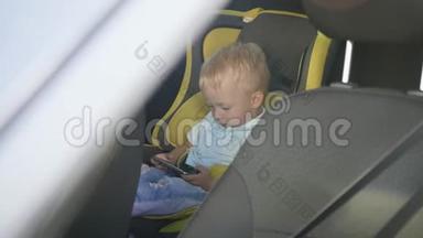 可爱的蹒跚学步的小男孩坐在汽车座椅上，从智能手机上观看视频。 小孩在车里玩智能手机
